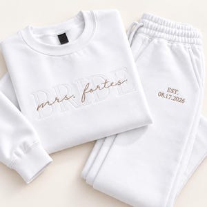 May include: A white long-sleeve sweatshirt and matching sweatpants set. The sweatshirt features the word BRIDE in large embossed letters with the text mrs. fortes embroidered in brown script. The sweatpants feature the text EST. 08.17.2026 in brown.