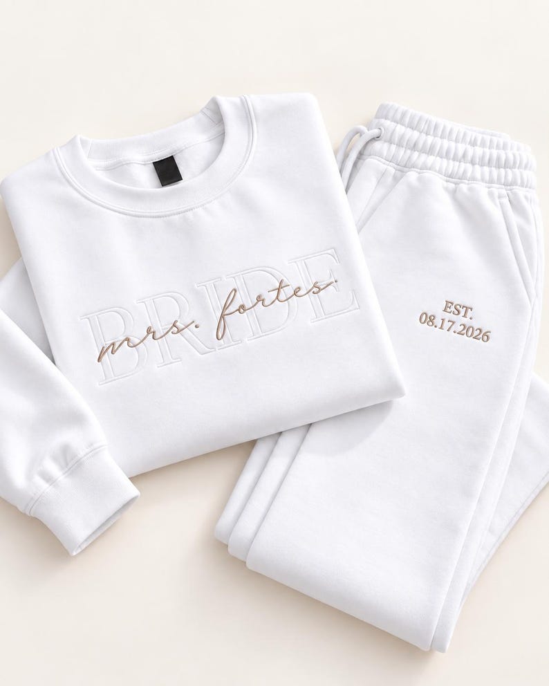 May include: A white long-sleeve sweatshirt and matching sweatpants set. The sweatshirt features the word BRIDE in large embossed letters with the text mrs. fortes embroidered in brown script. The sweatpants feature the text EST. 08.17.2026 in brown.