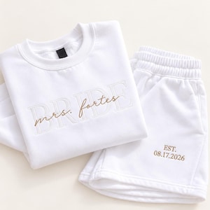May include: A white long-sleeve sweatshirt and matching shorts set. The sweatshirt features the word BRIDE in large white block letters with the text mrs. fortes embroidered in brown script. The shorts feature the text EST. 08.17.2026 in brown.