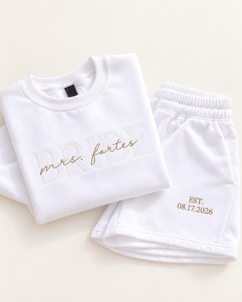 May include: A white long-sleeve sweatshirt and matching shorts set. The sweatshirt features the word BRIDE in large white block letters with the text mrs. fortes embroidered in brown script. The shorts feature the text EST. 08.17.2026 in brown.