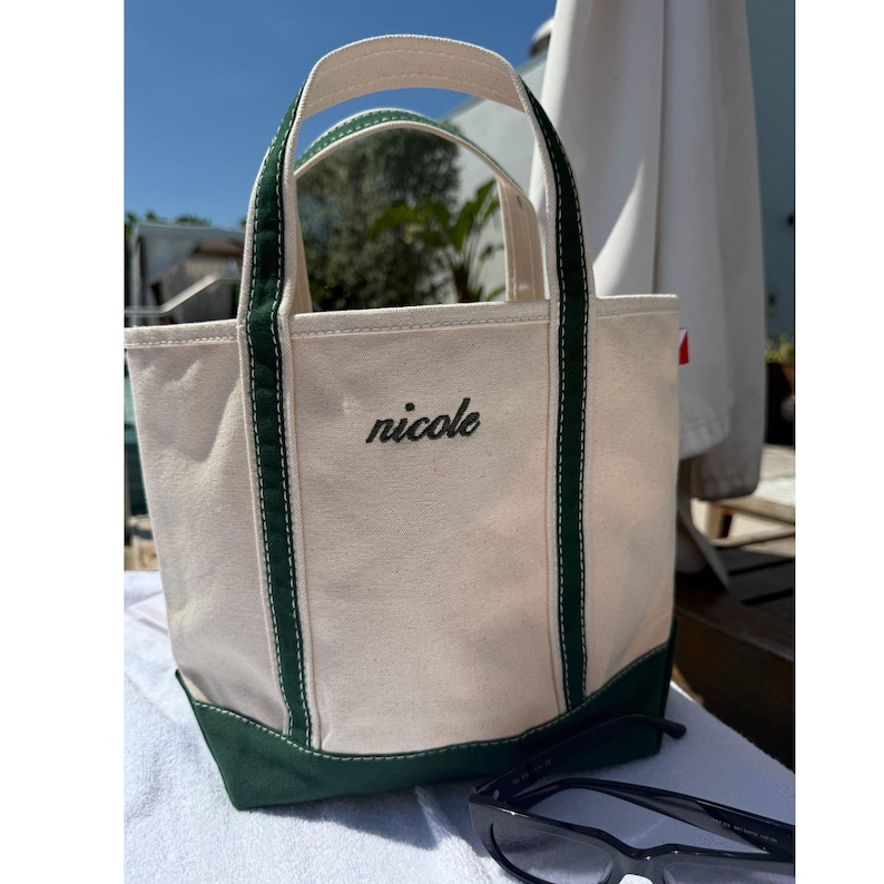 May include: A canvas tote bag with a cream-colored body and green accents. The bag features green handles and a green base. The name "nicole" is embroidered in black script on the front. Sunglasses rest in front of the bag.