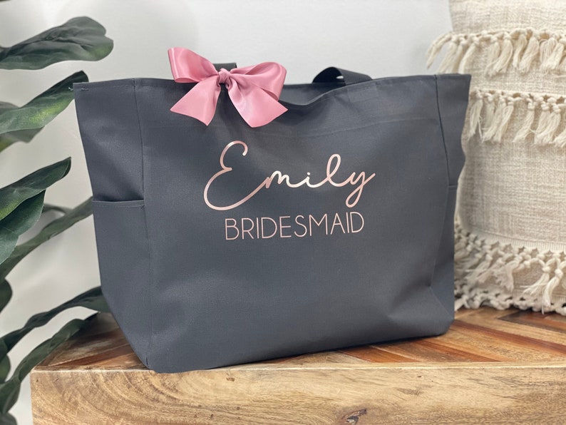 May include: A gray canvas tote bag with a pink bow and the text "Emily Bridesmaid" in rose gold lettering.
