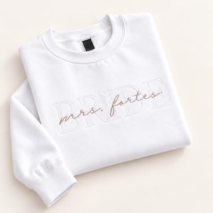 May include: A white long-sleeve crewneck sweatshirt featuring the word BRIDE embroidered in a large, tonal block font across the chest, with the text mrs. fortes stitched in a smaller, brown cursive script layered over the center.