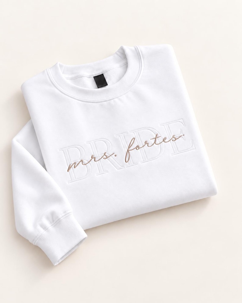 May include: A white long-sleeve crewneck sweatshirt featuring the word BRIDE embroidered in a large, tonal block font across the chest, with the text mrs. fortes stitched in a smaller, brown cursive script layered over the center.