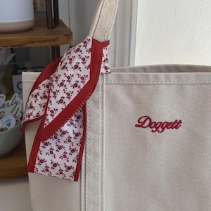 May include: A beige canvas tote bag featuring the embroidered red script text Doggett. A white fabric ribbon with a small red floral pattern and a solid red border is tied around one of the beige canvas handles of the tote bag.