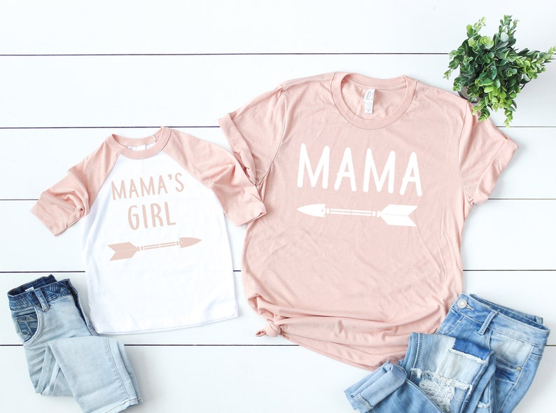 Pu&ograve; includere: Un set di due t-shirt, una rosa e una bianca con maniche rosa. La maglietta rosa dice "MAMA" con una freccia bianca sotto. La maglietta bianca dice "MAMA'S GIRL" con una freccia rosa sotto.