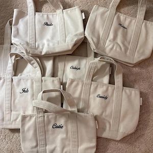 May include: A collection of six off-white canvas tote bags, each featuring two sturdy handles and black embroidered names including Paula, Kristen, Caleigh, Juls, Cassidy, and Catie. The bags are arranged in a group, showcasing their structured design.
