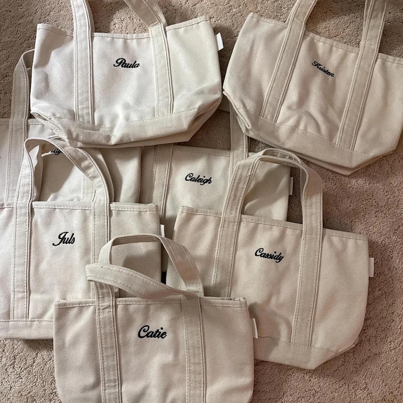 May include: A collection of six off-white canvas tote bags, each featuring two sturdy handles and black embroidered names including Paula, Kristen, Caleigh, Juls, Cassidy, and Catie. The bags are arranged in a group, showcasing their structured design.