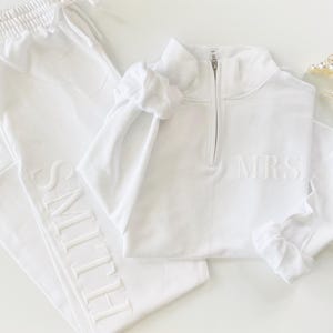 Op de afbeelding: Witte sweatpants met het woord "SMITH" geborduurd aan de zijkant en een wit sweatshirt met een halve ritssluiting en het woord "MRS" geborduurd op de voorkant.