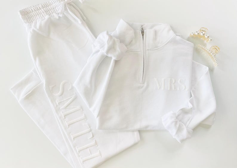 Op de afbeelding: Witte sweatpants met het woord "SMITH" geborduurd aan de zijkant en een wit sweatshirt met een halve ritssluiting en het woord "MRS" geborduurd op de voorkant.