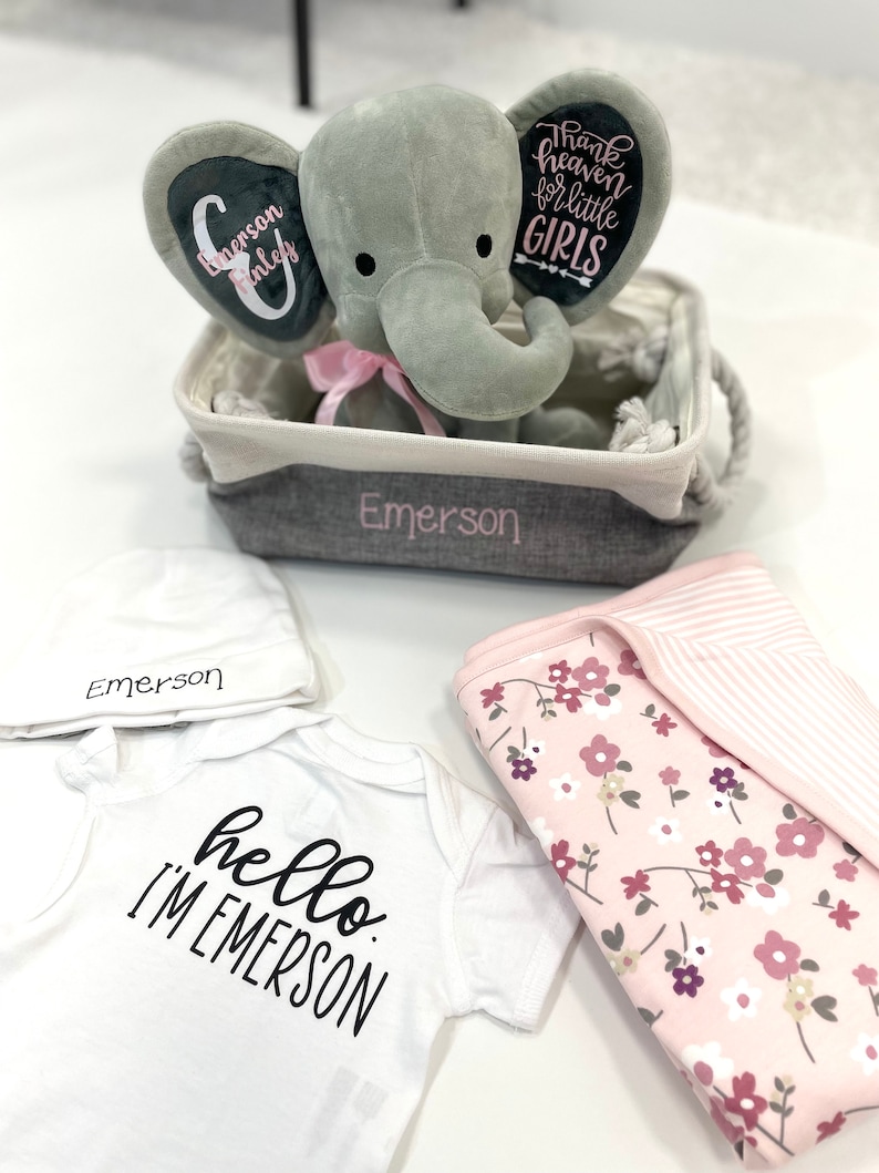 May include: A baby gift set featuring a gray plush elephant in a basket, a white onesie with "hello I'm Emerson" text, a white hat, and a pink floral blanket. The elephant has pink accents and the basket is embroidered with "Emerson".