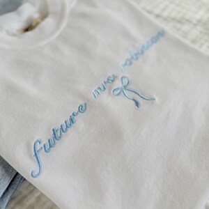 Op de afbeelding: Wit T-shirt met blauwe geborduurde tekst die "future mrs. Robinson" zegt met een blauwe strik.