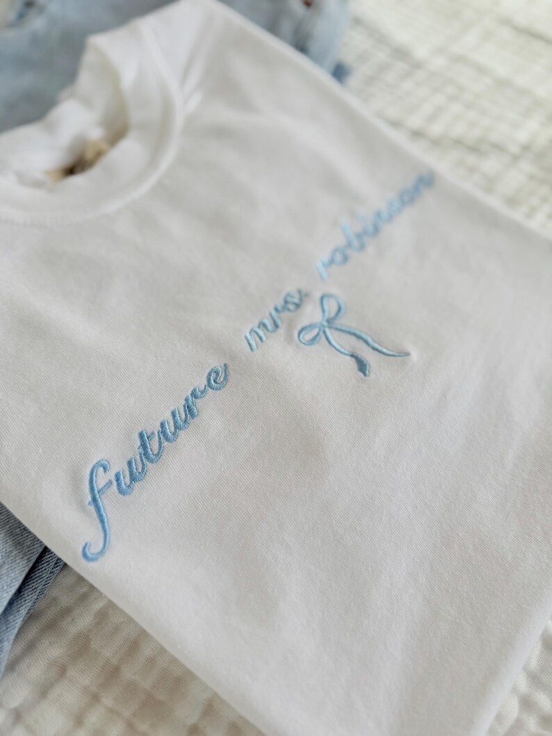 Op de afbeelding: Wit T-shirt met blauwe geborduurde tekst die "future mrs. Robinson" zegt met een blauwe strik.