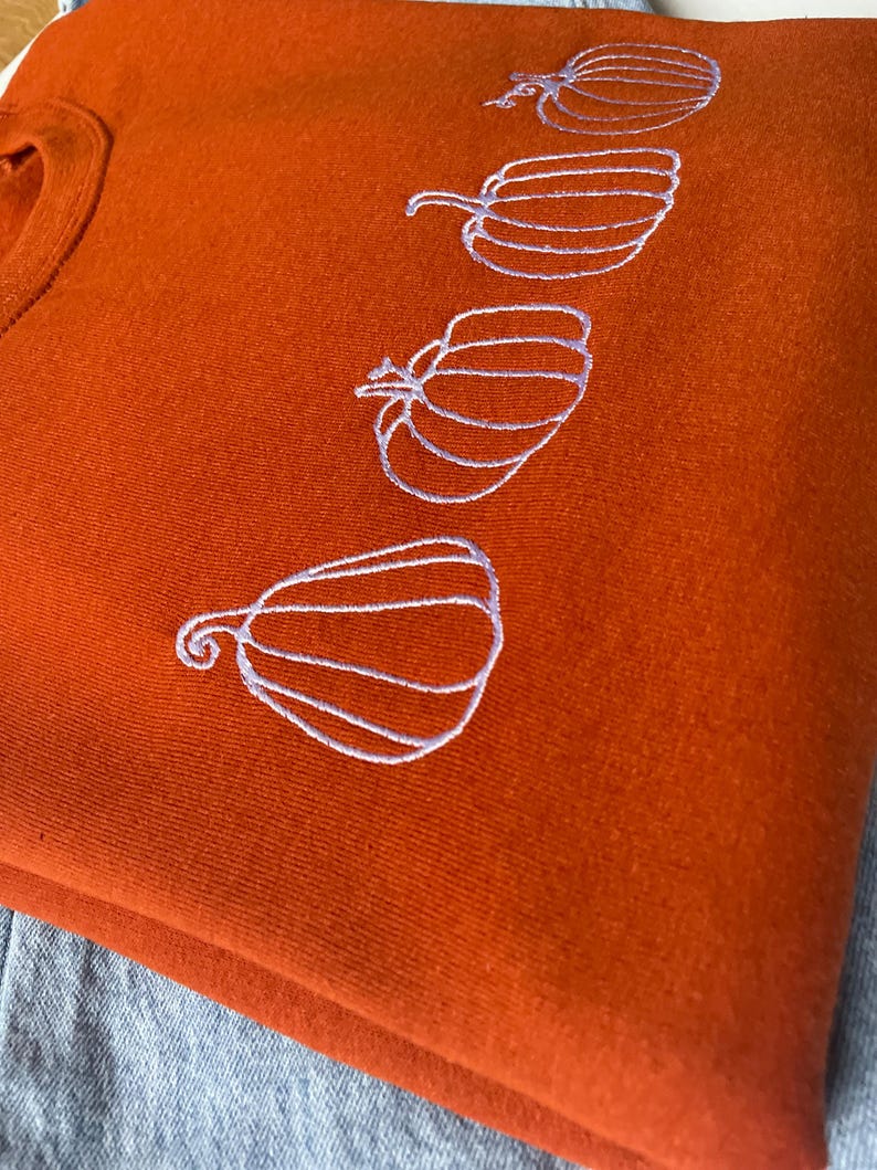 Puede incluir: Una sudadera de color &oacute;xido con un dise&ntilde;o bordado blanco de cuatro calabazas.