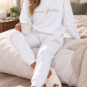 May include: A white crewneck sweatshirt featuring the text "BRIDE" in a light outline font with "mrs. fortes" in cursive script, paired with matching white sweatpants embroidered with "EST. 08.17.2026" in a light brown thread color.