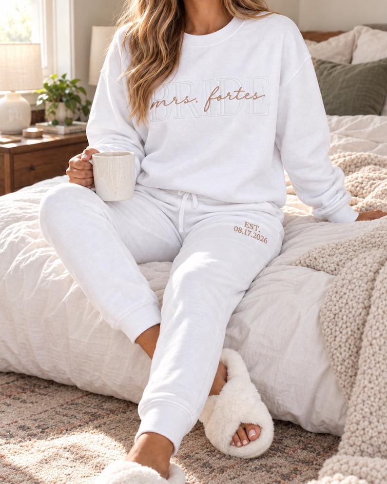 May include: A white crewneck sweatshirt featuring the text "BRIDE" in a light outline font with "mrs. fortes" in cursive script, paired with matching white sweatpants embroidered with "EST. 08.17.2026" in a light brown thread color.