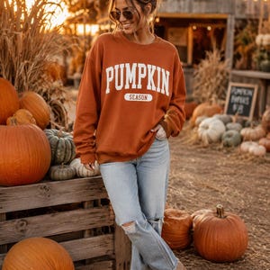Op de afbeelding: Een donkeroranje sweatshirt met lange mouwen en ronde hals, voorzien van de witte tekst PUMPKIN SEASON in een collegiaal lettertype op de borst. Het sweatshirt wordt gecombineerd met een lichtblauwe spijkerbroek met gescheurde kniedetails.