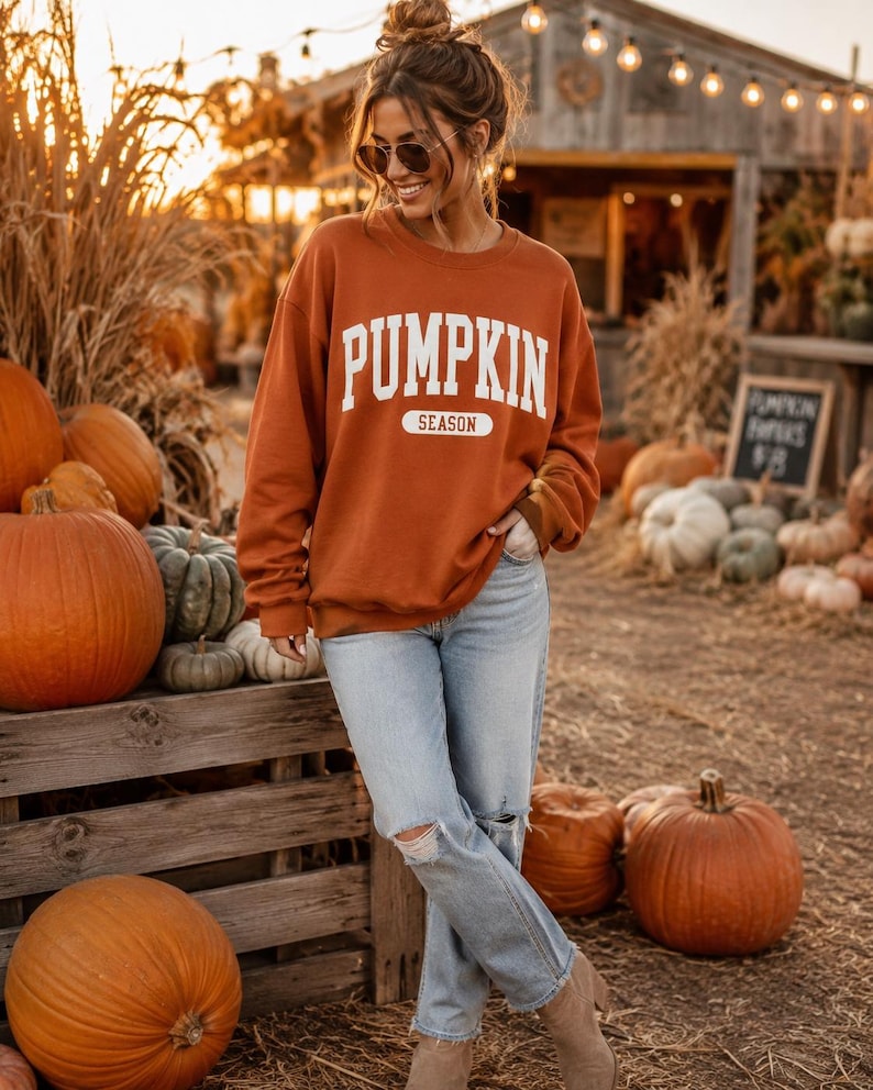 Op de afbeelding: Een donkeroranje sweatshirt met lange mouwen en ronde hals, voorzien van de witte tekst PUMPKIN SEASON in een collegiaal lettertype op de borst. Het sweatshirt wordt gecombineerd met een lichtblauwe spijkerbroek met gescheurde kniedetails.
