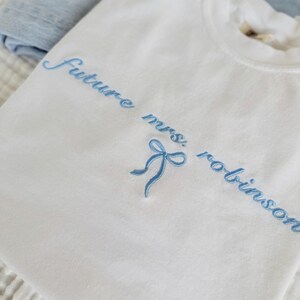 Op de afbeelding: Wit T-shirt met blauwe borduurwerk dat "Future Mrs. Robinson" zegt met een blauwe strik.