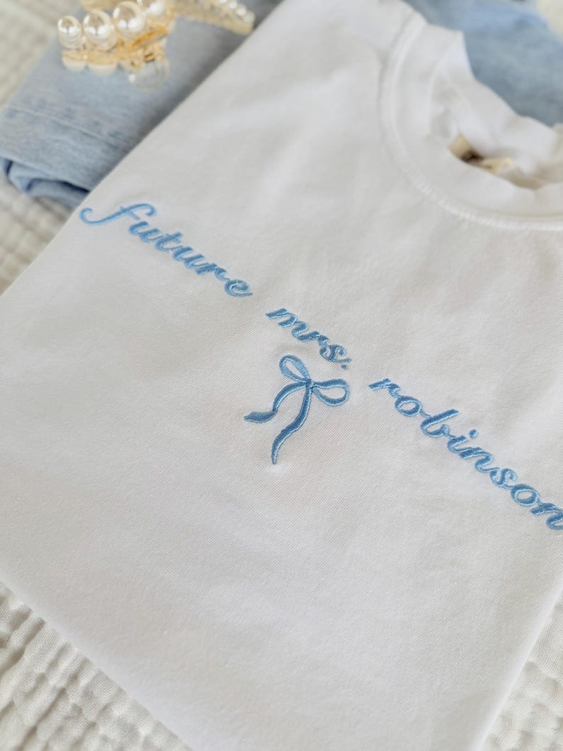 Op de afbeelding: Wit T-shirt met blauwe borduurwerk dat "Future Mrs. Robinson" zegt met een blauwe strik.