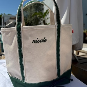 May include: A canvas tote bag with a cream-colored body, green base, and green handles. The name "nicole" is embroidered in black script on the front. Black sunglasses rest in front of the bag.