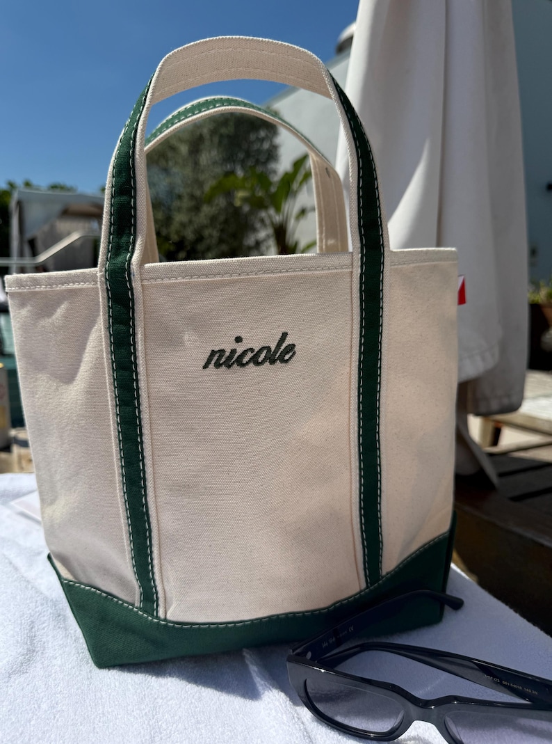 May include: A canvas tote bag with a cream-colored body, green base, and green handles. The name "nicole" is embroidered in black script on the front. Black sunglasses rest in front of the bag.
