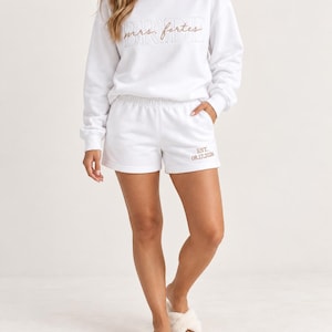 May include: A white long-sleeve crewneck sweatshirt and matching shorts set. The sweatshirt features the word BRIDE in white outline text with the script text mrs. fortes embroidered in tan. The shorts feature the text EST. 08.17.2026 in tan.