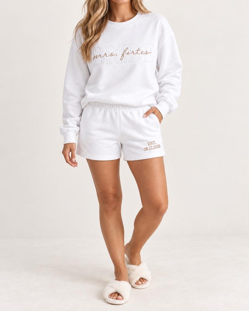 May include: A white long-sleeve crewneck sweatshirt and matching shorts set. The sweatshirt features the word BRIDE in white outline text with the script text mrs. fortes embroidered in tan. The shorts feature the text EST. 08.17.2026 in tan.