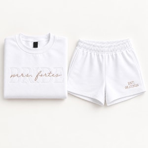 May include: A white sweatshirt and matching shorts set. The sweatshirt features the word BRIDE in large white block letters with the text mrs. fortes in brown script overlaid. The shorts feature the text EST. 08.17.2026 in brown embroidery.