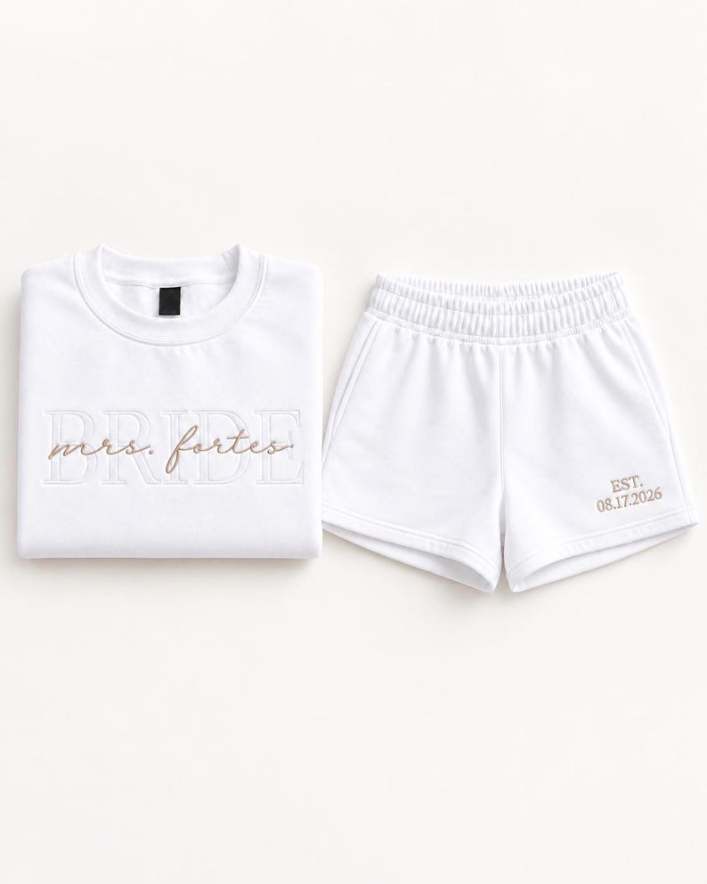 May include: A white sweatshirt and matching shorts set. The sweatshirt features the word BRIDE in large white block letters with the text mrs. fortes in brown script overlaid. The shorts feature the text EST. 08.17.2026 in brown embroidery.