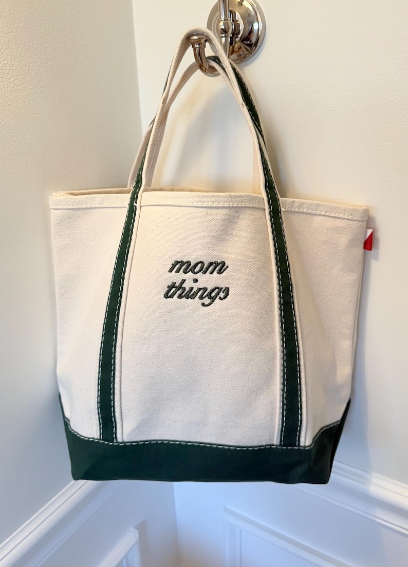 May include: Cream-colored canvas tote bag with dark green accents. The bag features a rectangular shape with a reinforced dark green base and handles. The words "mom things" are embroidered in black on the front.