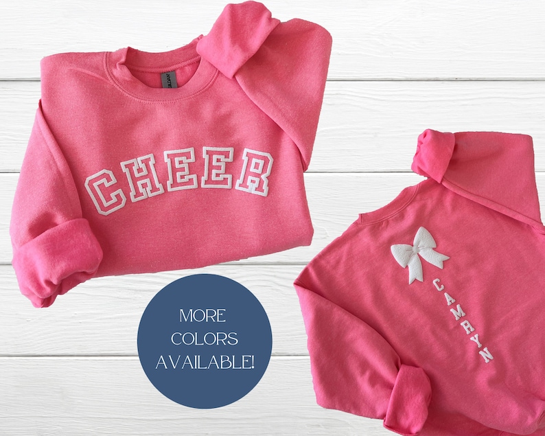 May include: Pink sweatshirt with the word "CHEER" in white embroidered letters on the front. The back of the sweatshirt has a white bow and the name "CAMRYN" embroidered in white letters.
