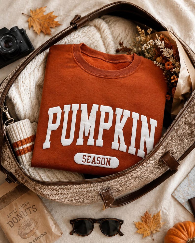 Op de afbeelding: Een roestkleurige sweater met ronde hals voorzien van de witte tekst PUMPKIN in een groot collegiaal lettertype en het woord SEASON op een kleinere witte rechthoekige patch eronder. Het kledingstuk is gevouwen in een bruine tas met leren hengsels.