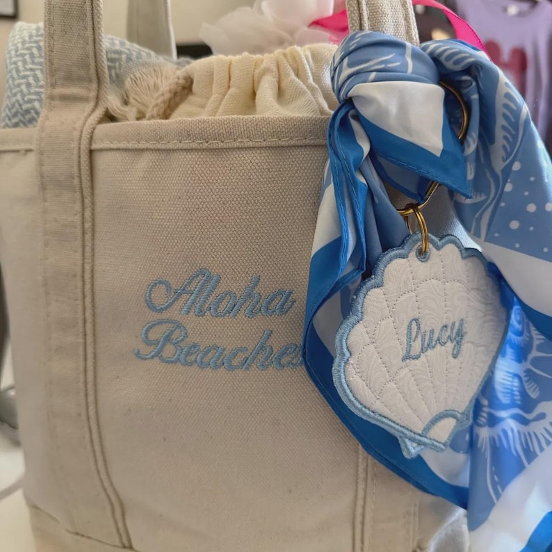 May include: A beige canvas tote bag featuring the embroidered text Aloha Beaches in light blue. A blue and white patterned scarf is tied to the handle, holding a white shell-shaped tag with the embroidered name Lucy in light blue thread.