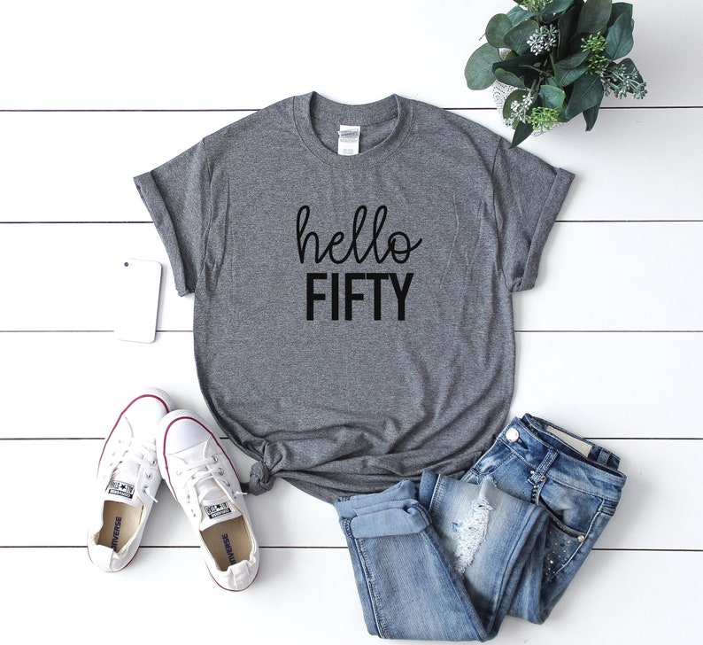 Pode incluir: Camiseta cinza com o texto "hello FIFTY" em letras pretas. A camiseta est&aacute; colocada sobre uma superf&iacute;cie de madeira branca com um par de t&ecirc;nis brancos, uma cal&ccedil;a jeans azul e uma planta em vaso.