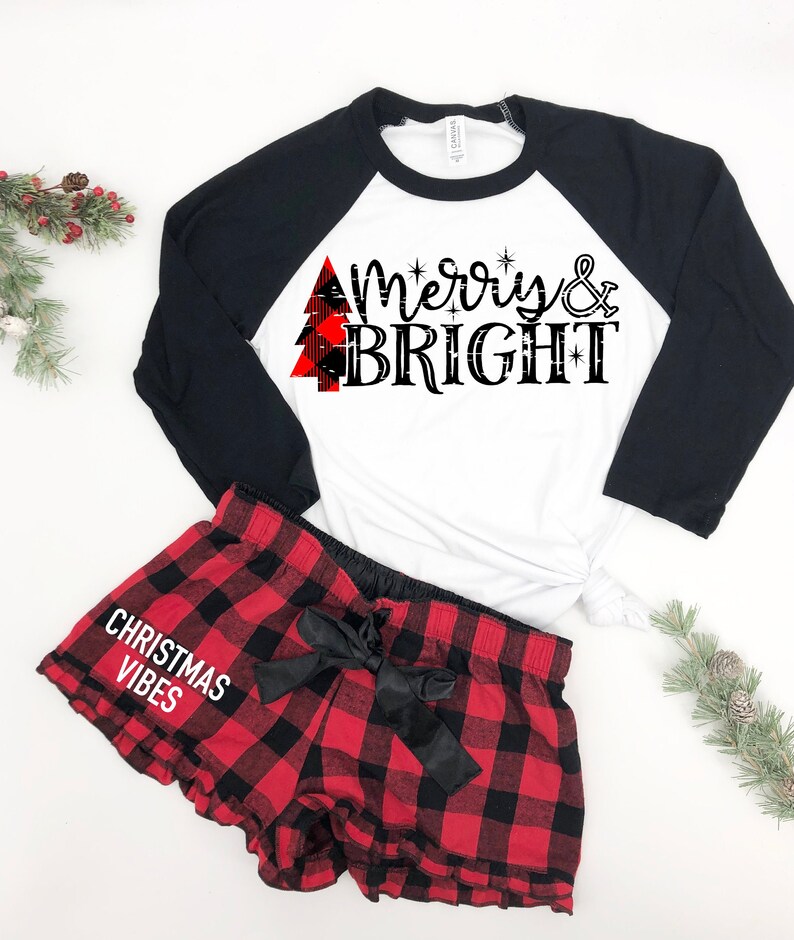 Pode incluir: Uma camiseta de beisebol branca e preta com uma &aacute;rvore de Natal xadrez vermelha e preta e o texto "Merry & Bright". Um short de pijama xadrez vermelho e preto com um la&ccedil;o de cetim preto e o texto "Christmas Vibes".