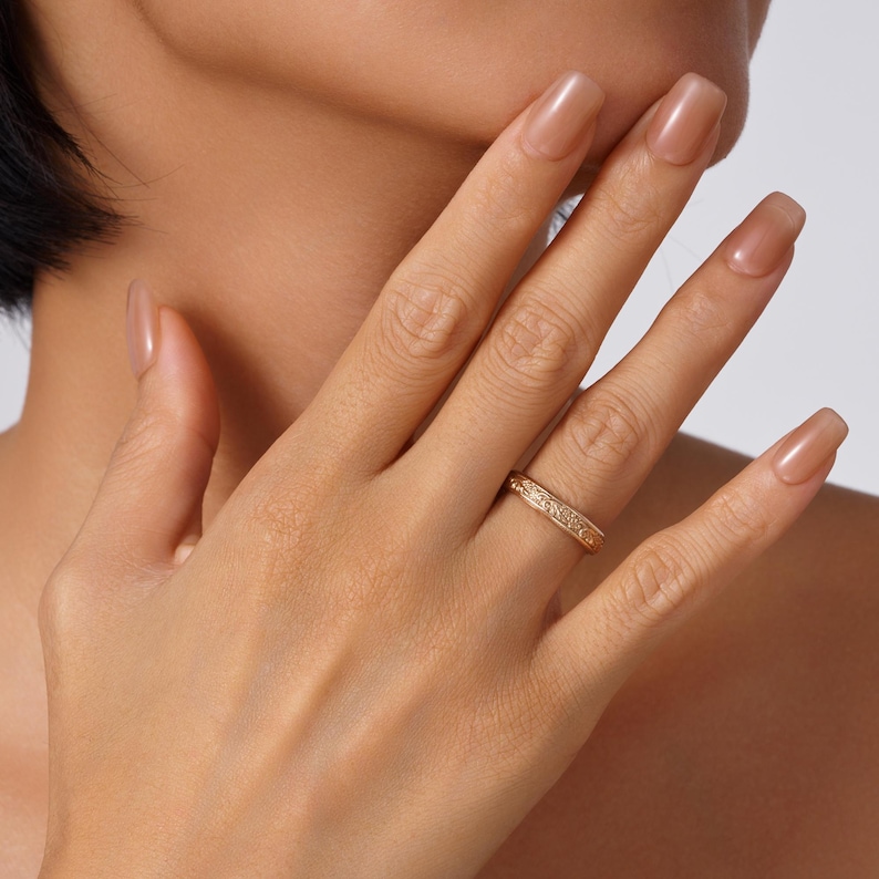 May include: A gold band ring featuring an intricate, embossed floral pattern along the entire outer surface. The ring has a polished finish and a consistent width, showcasing detailed craftsmanship with small, repeating engraved motifs around the band.