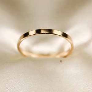 May include: A gold ring with a flat band. The ring is set against a soft, off-white background, creating a simple and elegant aesthetic. The ring's smooth surface reflects light, highlighting its metallic sheen.