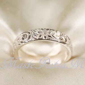 Puede incluir: Un anillo de plata con un patrón floral grabado. El anillo presenta un diseño floral central rodeado de grabados detallados y arremolinados. El anillo se encuentra sobre un fondo suave y blanquecino, resaltando sus intrincados detalles.