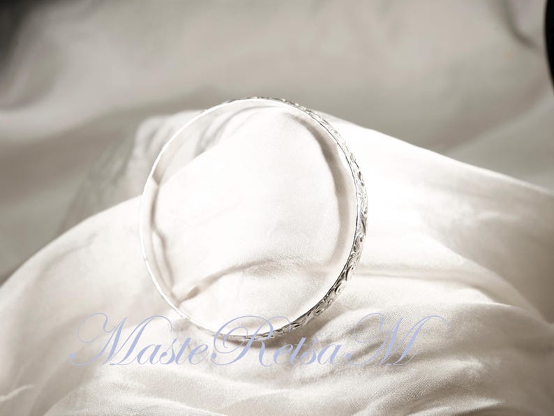 May include: A silver bangle bracelet with a delicate, intricate design. The bracelet is smooth and shiny, and it appears to be made of high-quality metal.