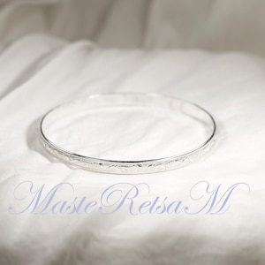 May include: A silver bangle bracelet with an engraved floral pattern. The bracelet is circular and rests on a white, textured surface. The image includes the text "Maste Retsa M" in a cursive font.