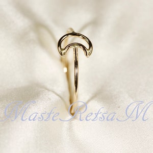 May include: A gold-colored ring featuring a crescent moon design. The ring has a slender band and a delicate, openwork moon shape. The ring is set against a soft, white background.