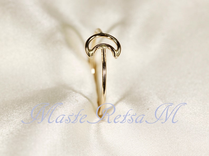 May include: A gold-colored ring featuring a crescent moon design. The ring has a slender band and a delicate, openwork moon shape. The ring is set against a soft, white background.