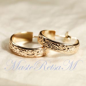 May include: A pair of gold-colored hoop earrings with intricate floral and scrollwork designs. The earrings are displayed on a soft, off-white surface. The text "Maste Retsa M" is visible in the lower portion of the image.