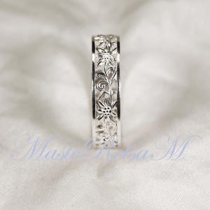 May include: A silver ring with a floral design. The ring features an intricate pattern of flowers and vines, creating a delicate and elegant look. The ring is set against a white background.