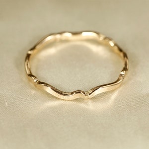 Könnte beinhalten: Ein goldfarbener Ring mit einem wellenförmigen, organischen Design. Der Ring hat eine strukturierte Oberfläche und eine durchgehende, fließende Form. Das Design des Rings erinnert an eine natürliche, fließende Form. Der Ring ist ein Schmuckstück.