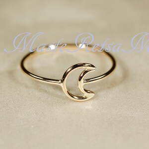 May include: A gold-colored ring featuring a crescent moon design. The ring is delicate and minimalist, with the moon shape as the central focus. The text "Maste Ressa M" is written in a light blue cursive font.