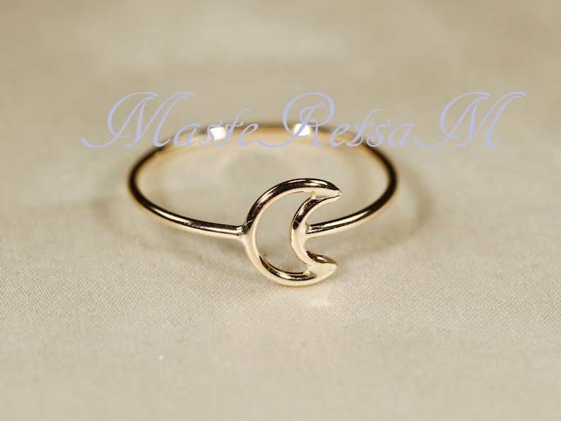 May include: A gold-colored ring featuring a crescent moon design. The ring is delicate and minimalist, with the moon shape as the central focus. The text "Maste Ressa M" is written in a light blue cursive font.