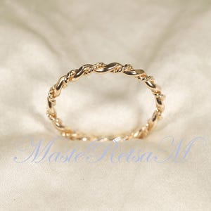 Puede incluir: Un anillo dorado con un diseño de cuerda trenzada. El anillo es circular y parece estar hecho de metal. El anillo está sobre un fondo suave y blanquecino. El diseño del anillo es intrincado y detallado.