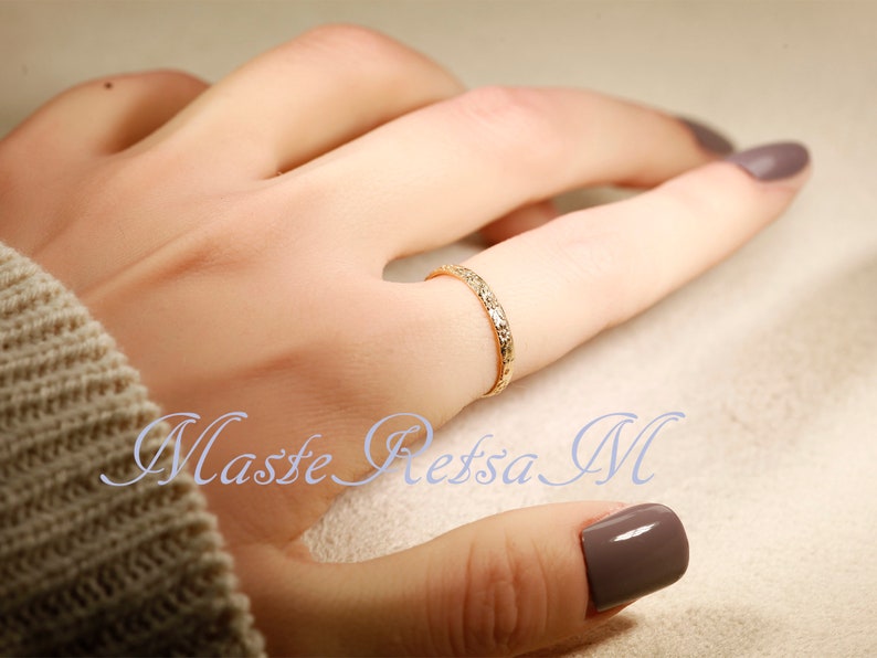 Puede incluir: Un delicado anillo de banda de oro con una superficie texturizada. El anillo se lleva en un dedo con un esmalte de u&ntilde;as marr&oacute;n claro.